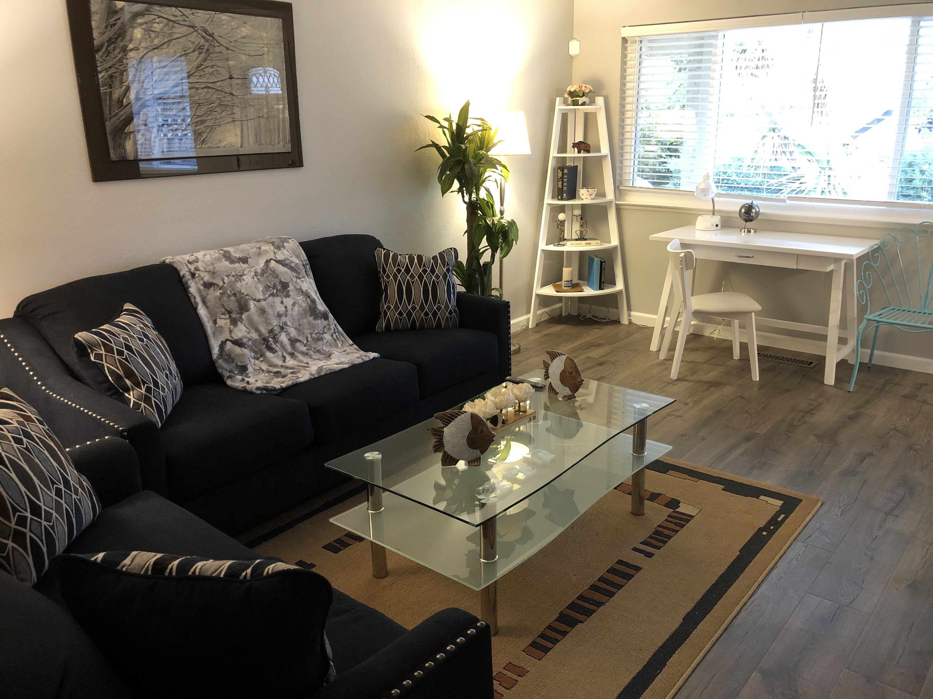 Ein Wohnzimmer mit einem dunkelgrauen Sofa, einem Glastisch, einem weißen Schreibtisch und einer Pflanze.