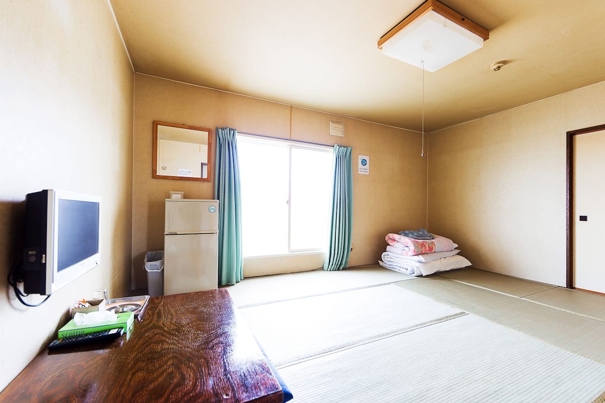 Une chambre d’hôtel avec un bureau en bois, une télévision, un réfrigérateur, une fenêtre avec des rideaux et une pile d’oreillers.