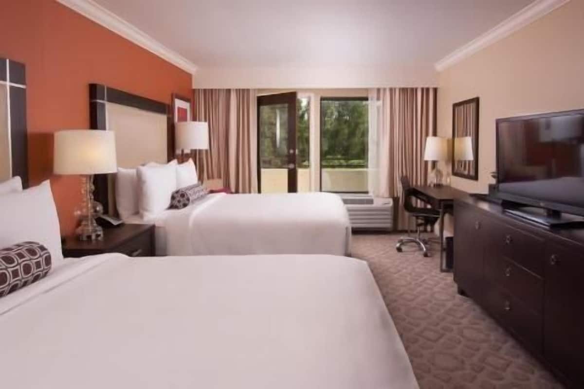 A hotel room with two beds, a desk, a television, and a view of the outdoors.