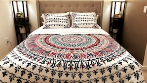 Un lit recouvert d’une couvre-lit à motifs, deux oreillers aux motifs similaires et deux tables de chevet avec des lampes et des plantes.