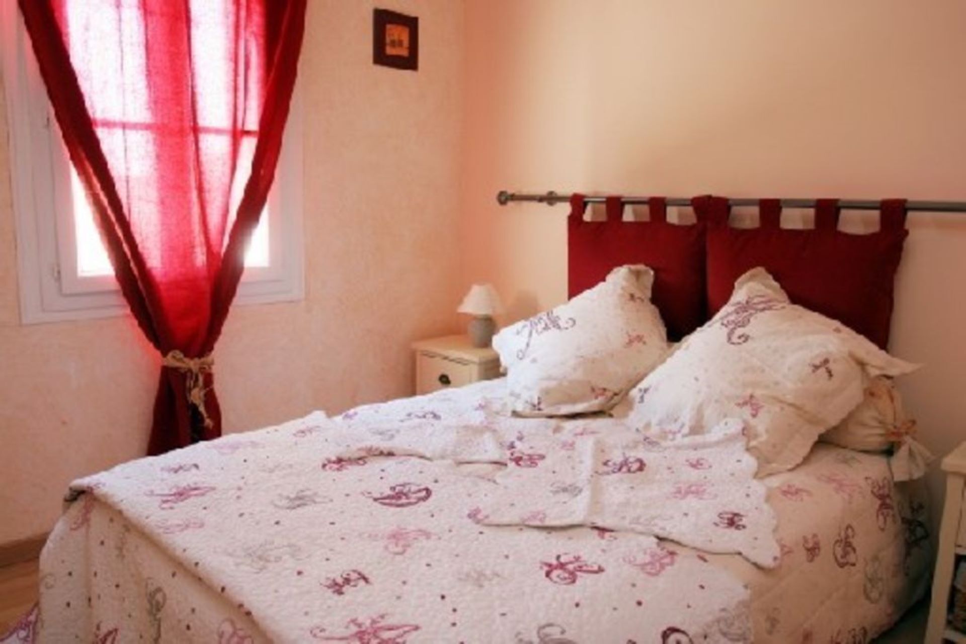 A bedroom with a bed, a nightstand, a lamp, and a window with red curtains.