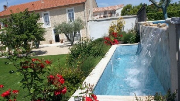 Una casa de piedra con techo de tejas, piscina con cascada y un jardín bien cuidado con flores rojas.
