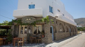 A two-story building with a stone facade and blue shutters. The ground floor has a sign reading "Hotel Tabernas Restaurant." There are outdoor tables and chairs, and a potted plant on the porch. - Louiza Hotel (Paros)