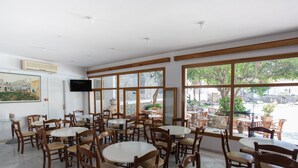 A restaurant with multiple round tables and wooden chairs, large windows with a view of the outside, and a flat-screen TV mounted on the wall. - Louiza Hotel (Paros)