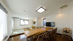 A spacious room with a long wooden table and chairs, a flat-screen TV mounted on the wall, and a window providing natural light. - Kamanoyado - Tennoji (Osaka)