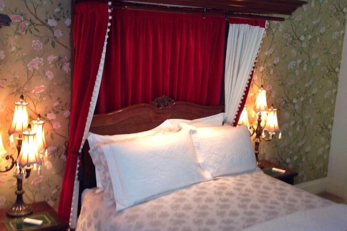 A four-poster bed with red and white drapes, two bedside lamps, and floral wallpaper.