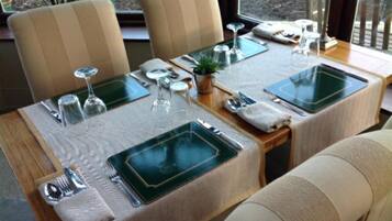 Mesa de comedor lista con manteles individuales verdes, cubiertos y cristalería, rodeada de sillas tapizadas en color beige.