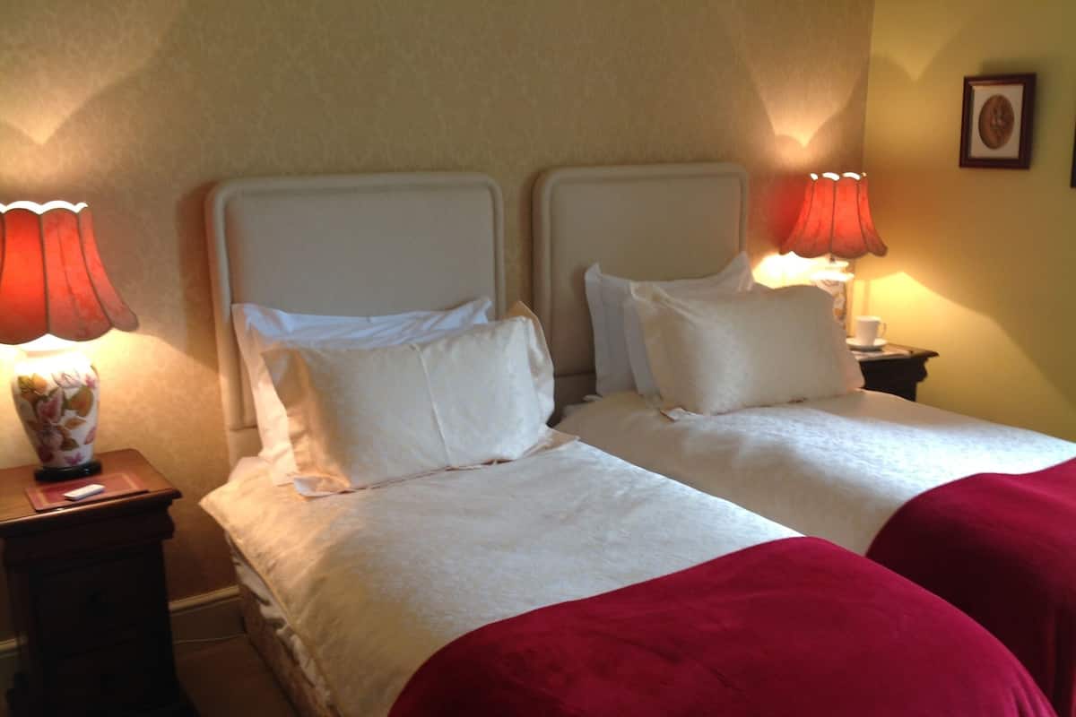 A hotel room with two beds, each with white linens and red throw blankets. There are two bedside tables with lamps and a framed picture on the wall.