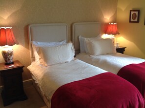 Habitación de hotel con dos camas, cada una con ropa blanca y mantas rojas. Dos mesitas de noche con lámparas y un cuadro en la pared.