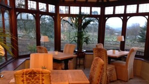 A conservatory with a wooden table and chairs, large windows, and a view of trees. - Westgate Manor (Bishop Auckland)