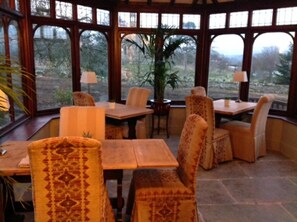 Un invernadero con una mesa y sillas de madera, ventanas amplias y vistas a árboles.