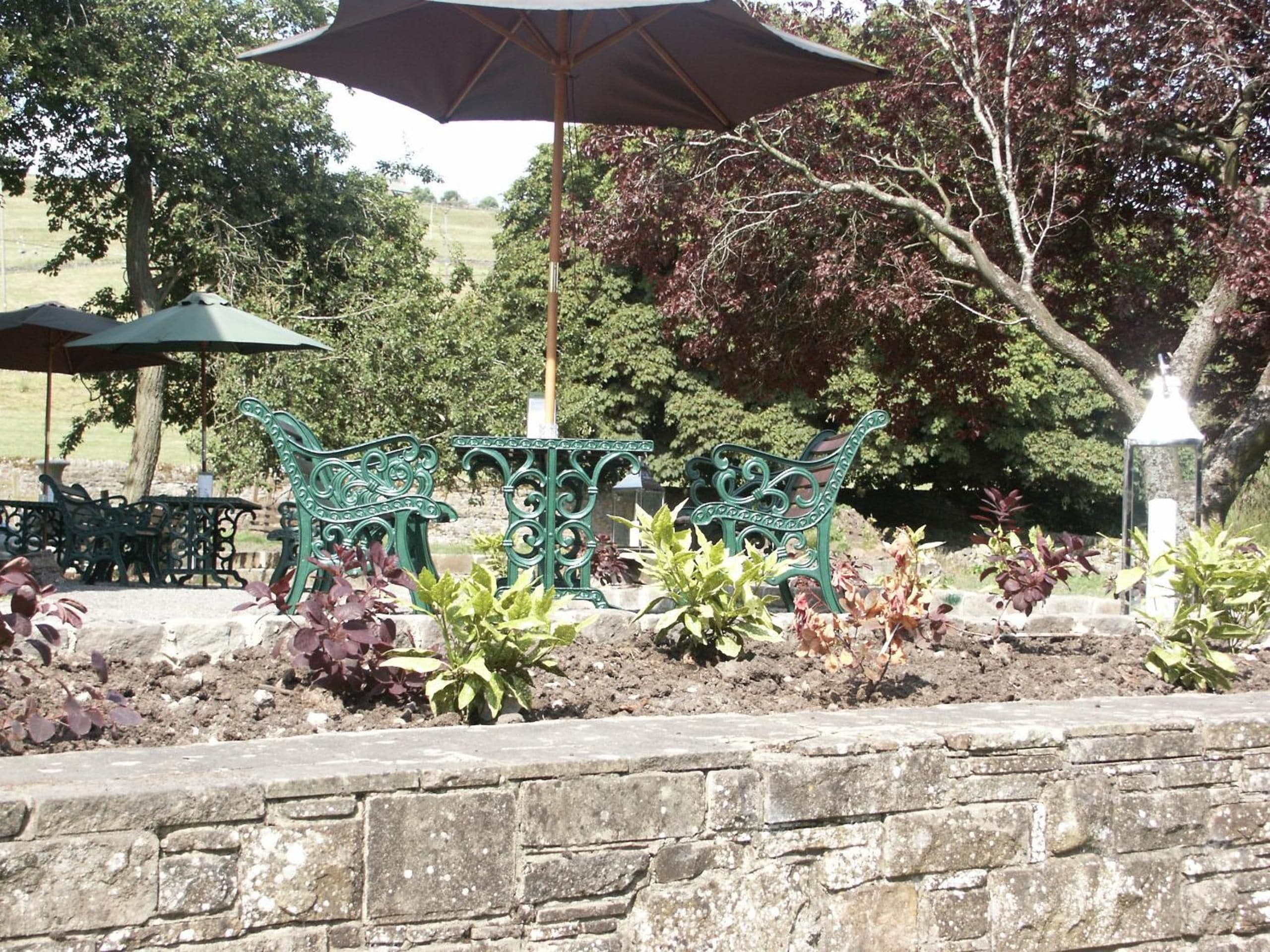 Un jardín con muro de piedra, banco de metal verde y dos sombrillas verdes.