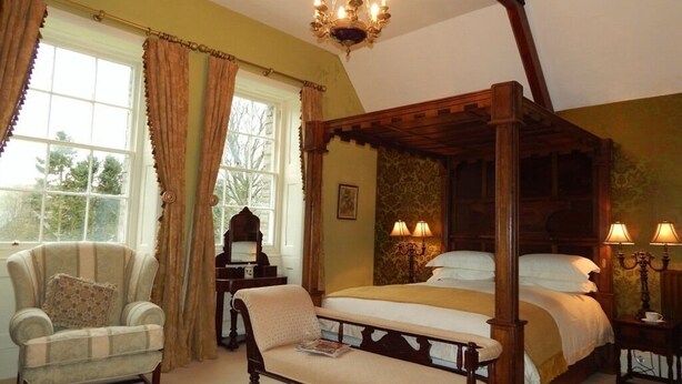 A four-poster bed with a wooden canopy, a sitting chair, a bedside table with lamps, and a window with curtains. - Westgate Manor (Bishop Auckland)