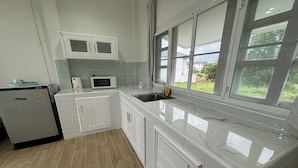 A kitchen with white cabinets, a microwave, a sink, and a window overlooking a garden. - Tann Anda Resort (Thep Krasattri)