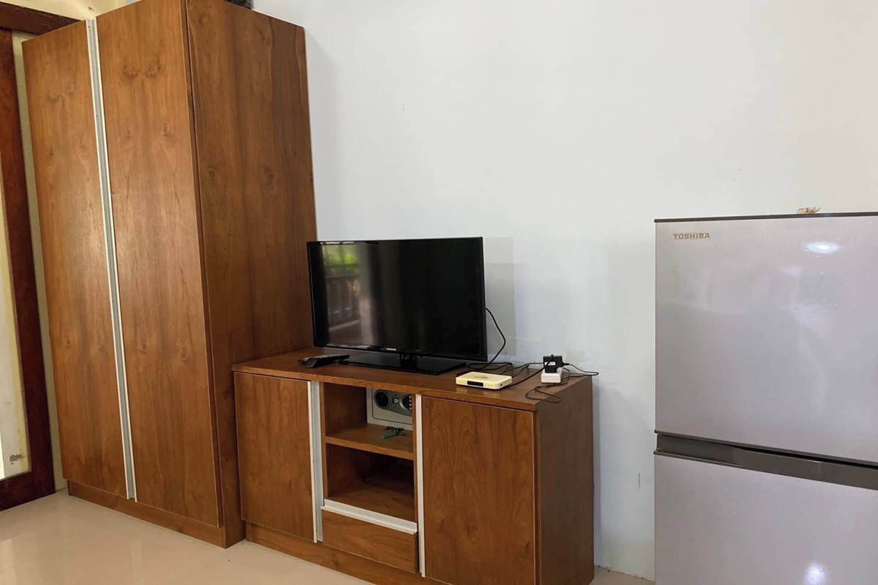 A room with a wooden wardrobe, a flat-screen TV on a wooden stand, and a white refrigerator.