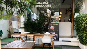 An outdoor seating area with wooden tables and chairs, a small garden with artificial grass, and a café entrance with a sign advertising 'All Day Breakfast'. - Two Gals And The Pig - Hostel (Chiang Mai)