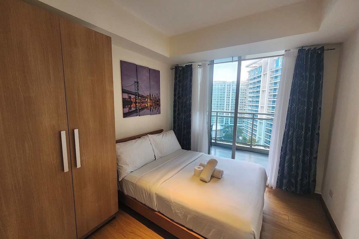A bedroom with a bed, a wooden wardrobe, a cityscape painting, and a large window with curtains.