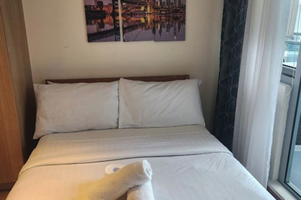 Habitación de hotel con una cama, dos almohadas, una ventana con cortinas y un cuadro de un paisaje urbano en la pared.