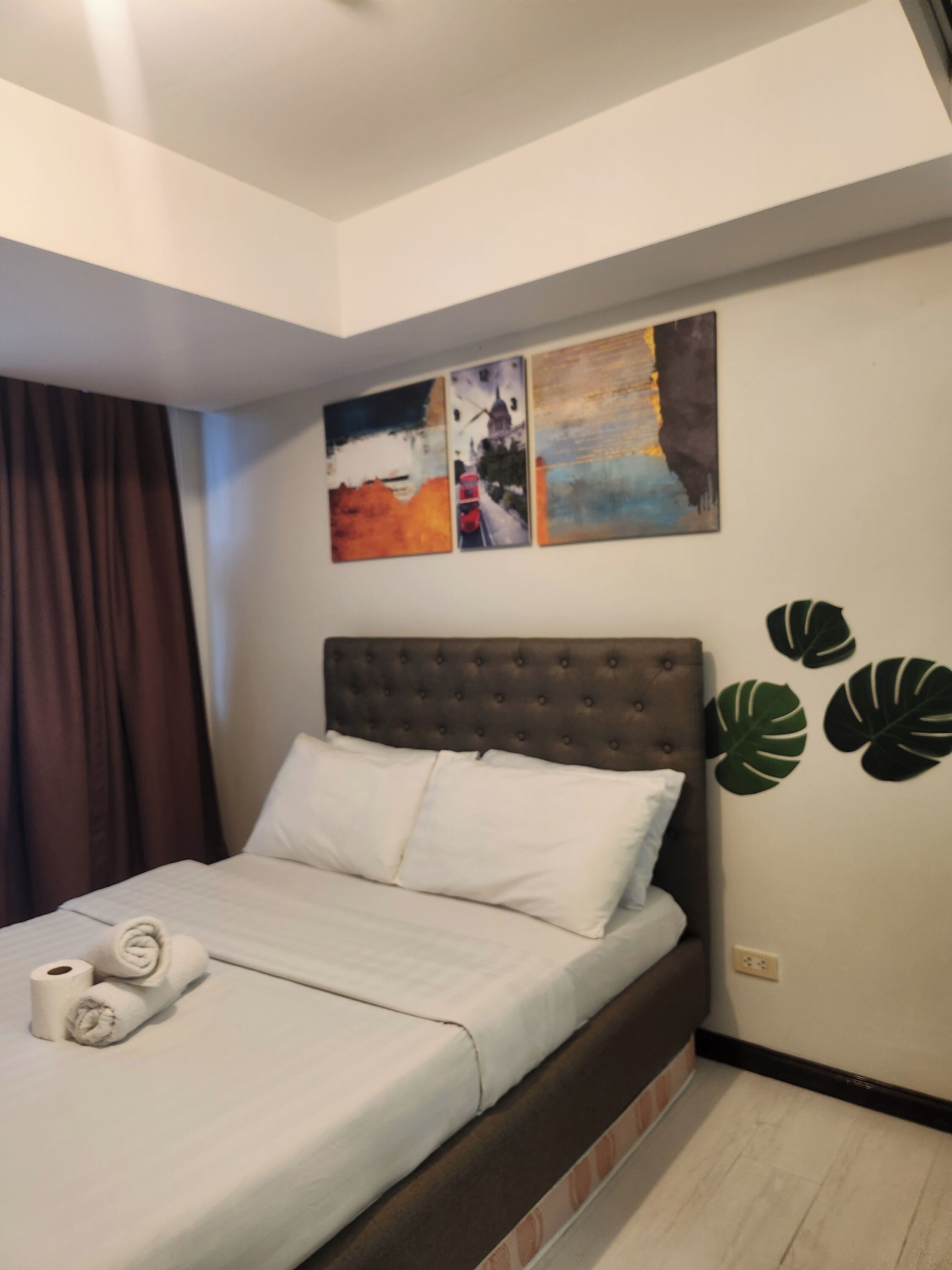 A neatly made bed with white linens and pillows, a tufted headboard, and a wall with abstract art and a plant decal.
