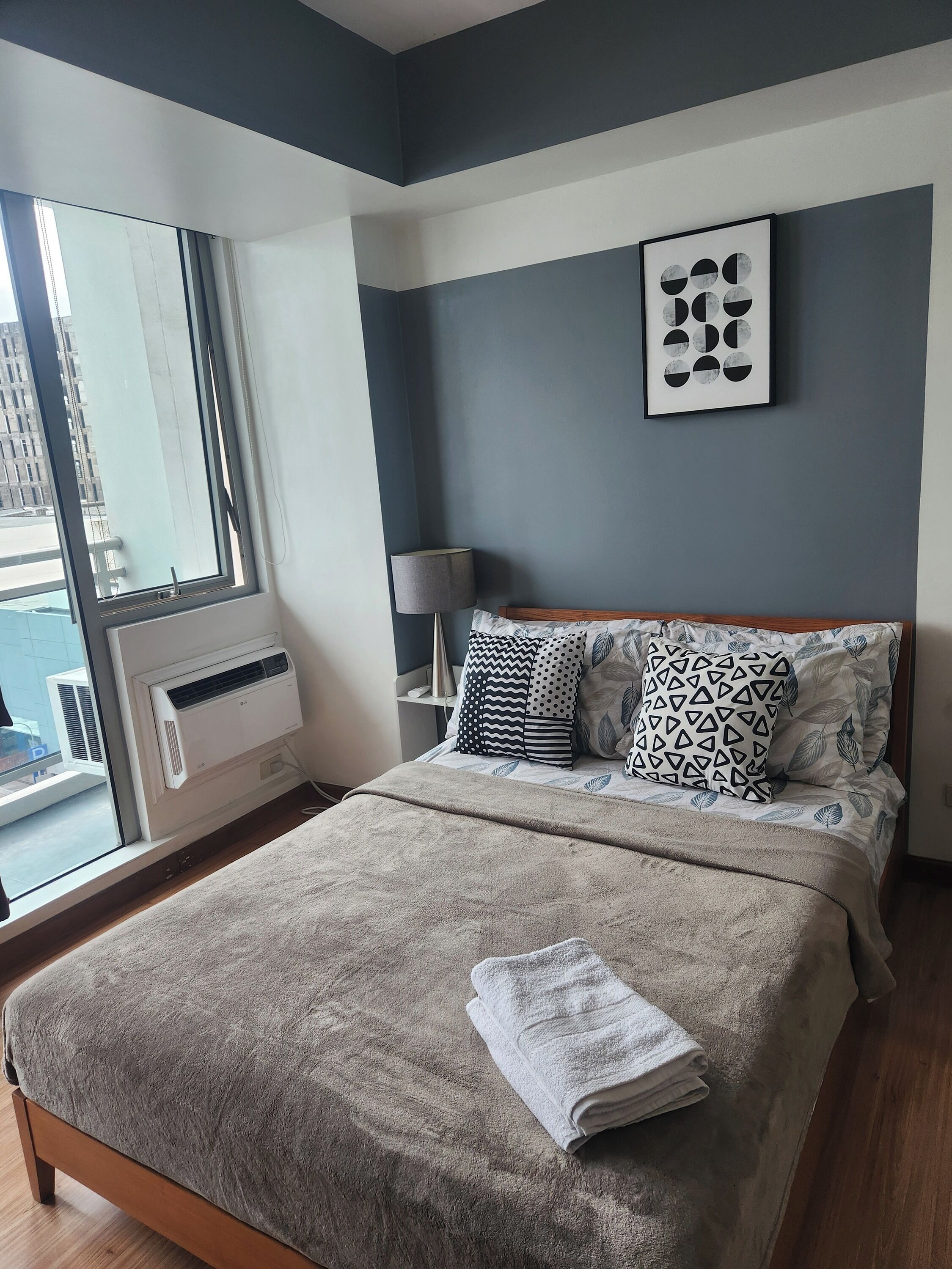 A bedroom with a large window, a bed with patterned pillows, a bedside lamp, and a wall-mounted artwork.