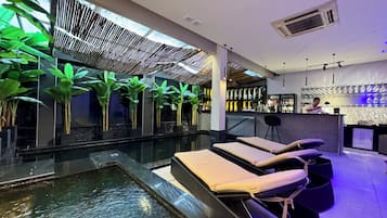 A modern indoor pool area with lounge chairs, a bar, and potted plants.