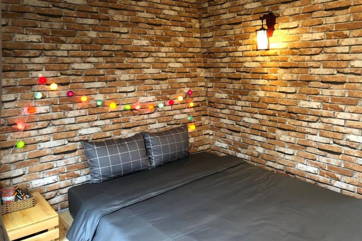 A room with a brick wall, a bed with grey bedding, and colorful string lights.