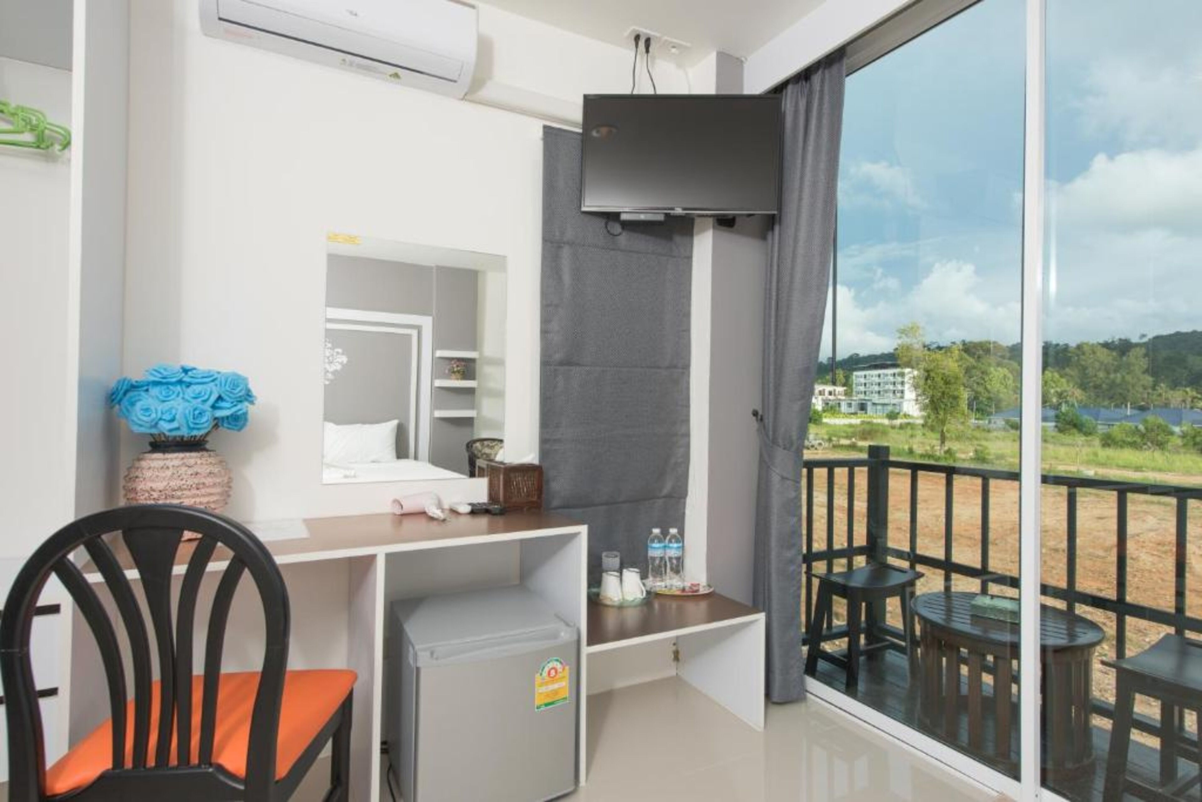 Een balkon met uitzicht op een meer en gebouwen, een klein bureau met stoel, een aan de muur bevestigde televisie en een koelkast.