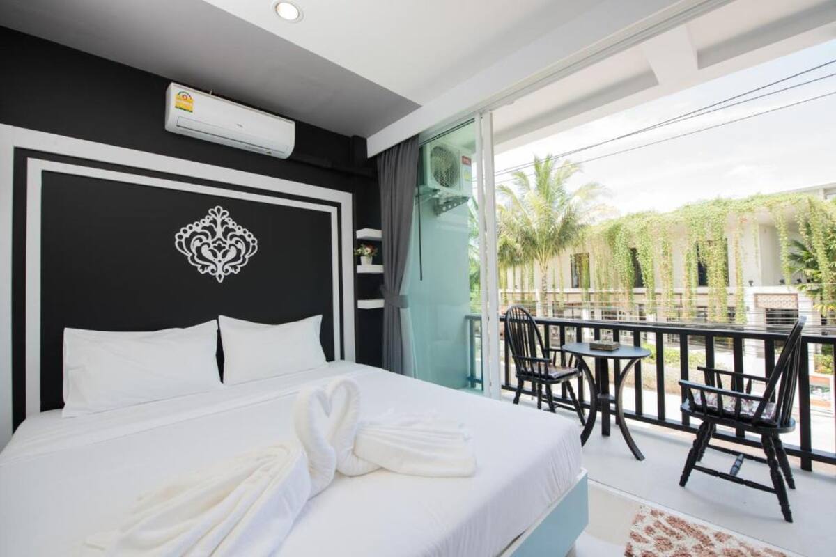 A hotel room with a large bed, a decorative wall piece, a balcony with chairs, and a view of palm trees.