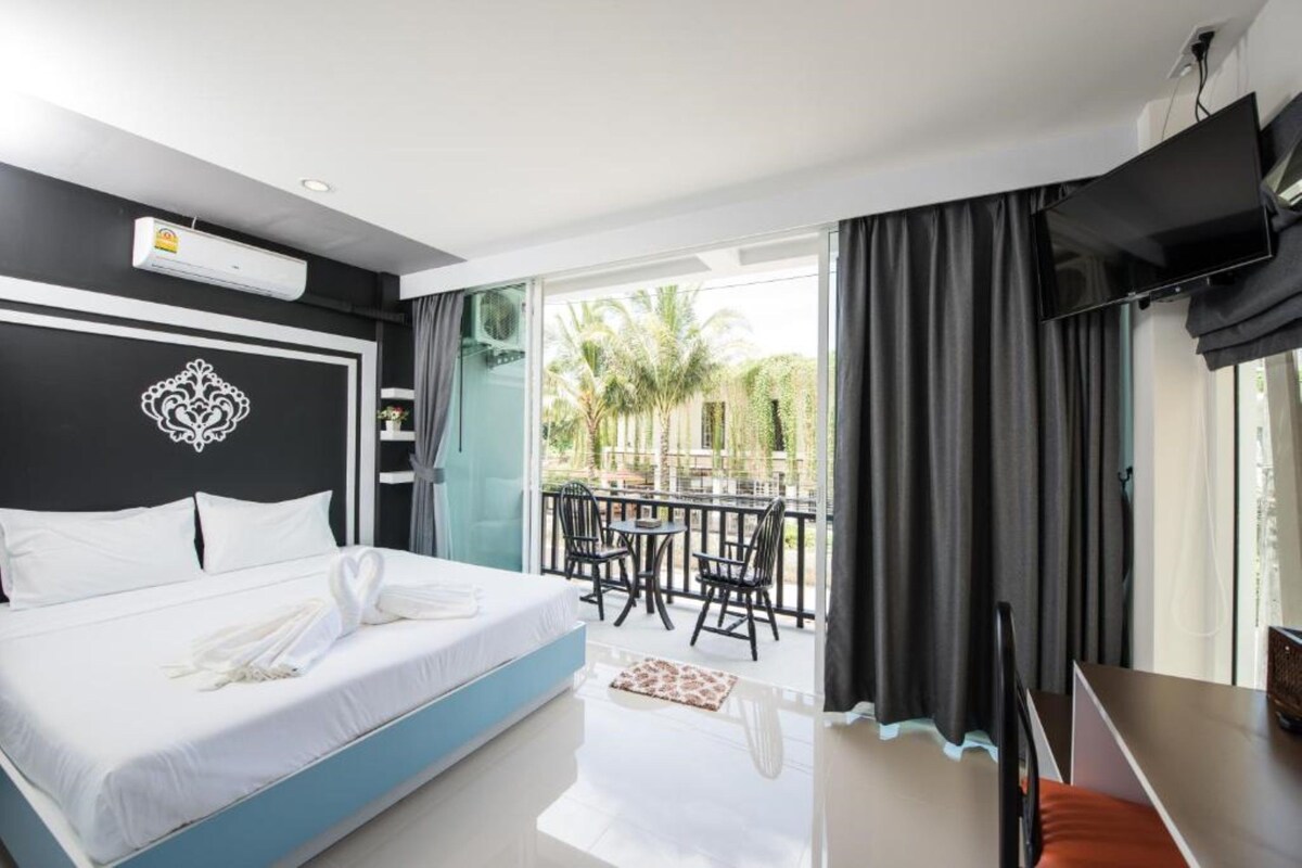 Habitación de hotel con cama, televisión, balcón con mesa y sillas, y vistas a palmeras.