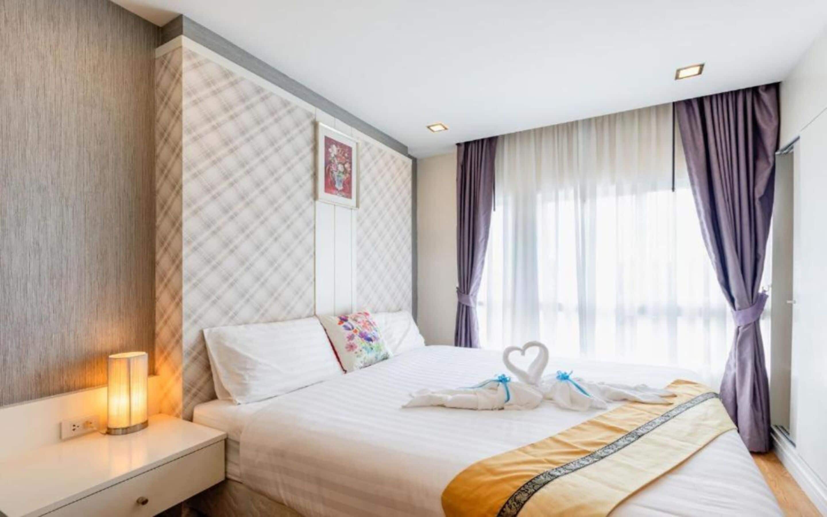 Un cuarto de hotel con una cama bien hecha, una lámpara de noche y un ventanal con cortinas.