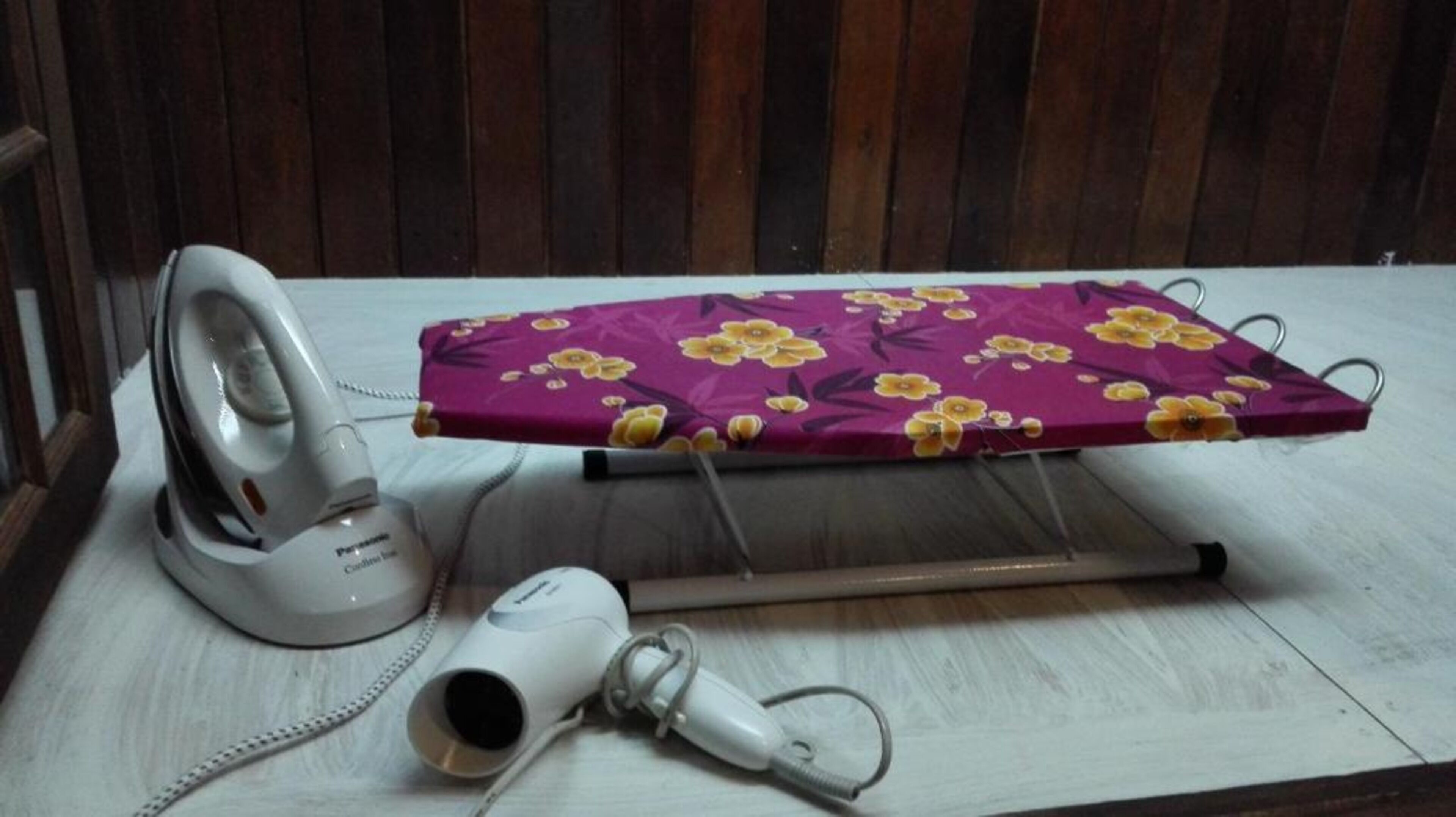 An ironing board with a floral cloth cover, an iron, and a hairdryer on a wooden surface.