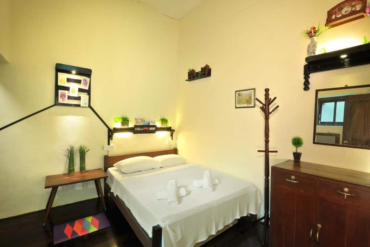 A neatly arranged hotel room with a bed, bedside table, dresser, and wall-mounted shelf with decorative items.