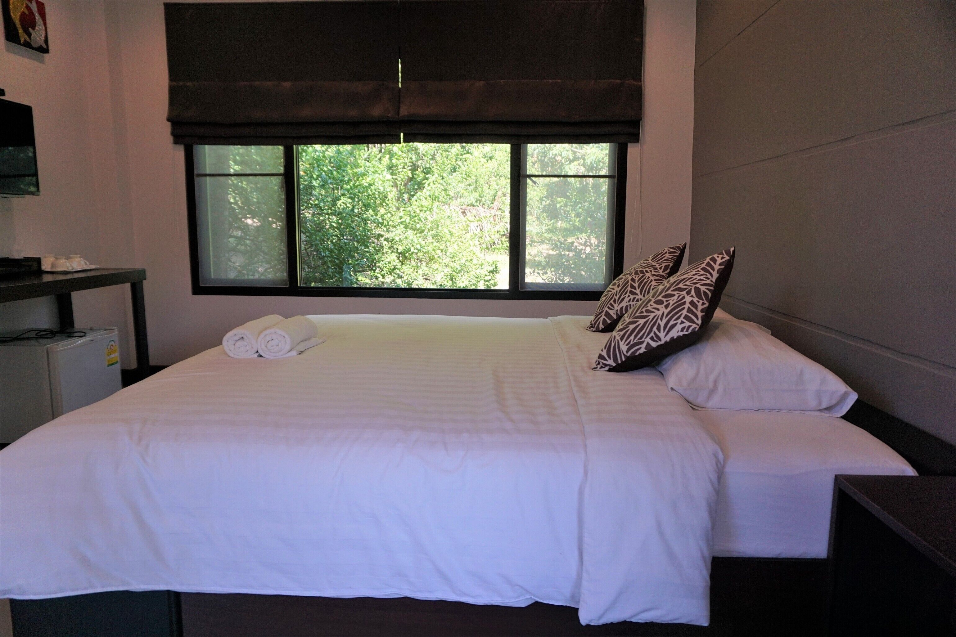 A neatly made bed with white linens and two rolled towels, a window with a view of greenery, and a dark-colored headboard.