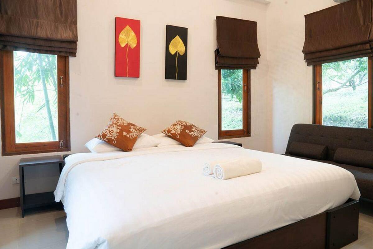 A bedroom with a bed, two pillows, a bedside table, and a chair. There are two windows with curtains, and artwork on the wall.