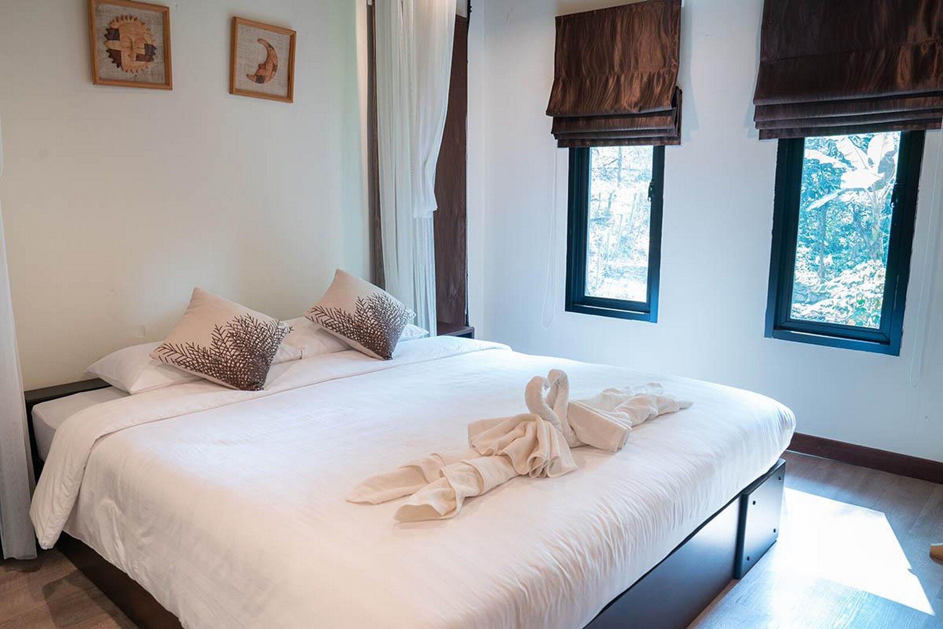 A neatly made bed with a towel and a pillow, a window with a view of trees, and two framed artworks on the wall.