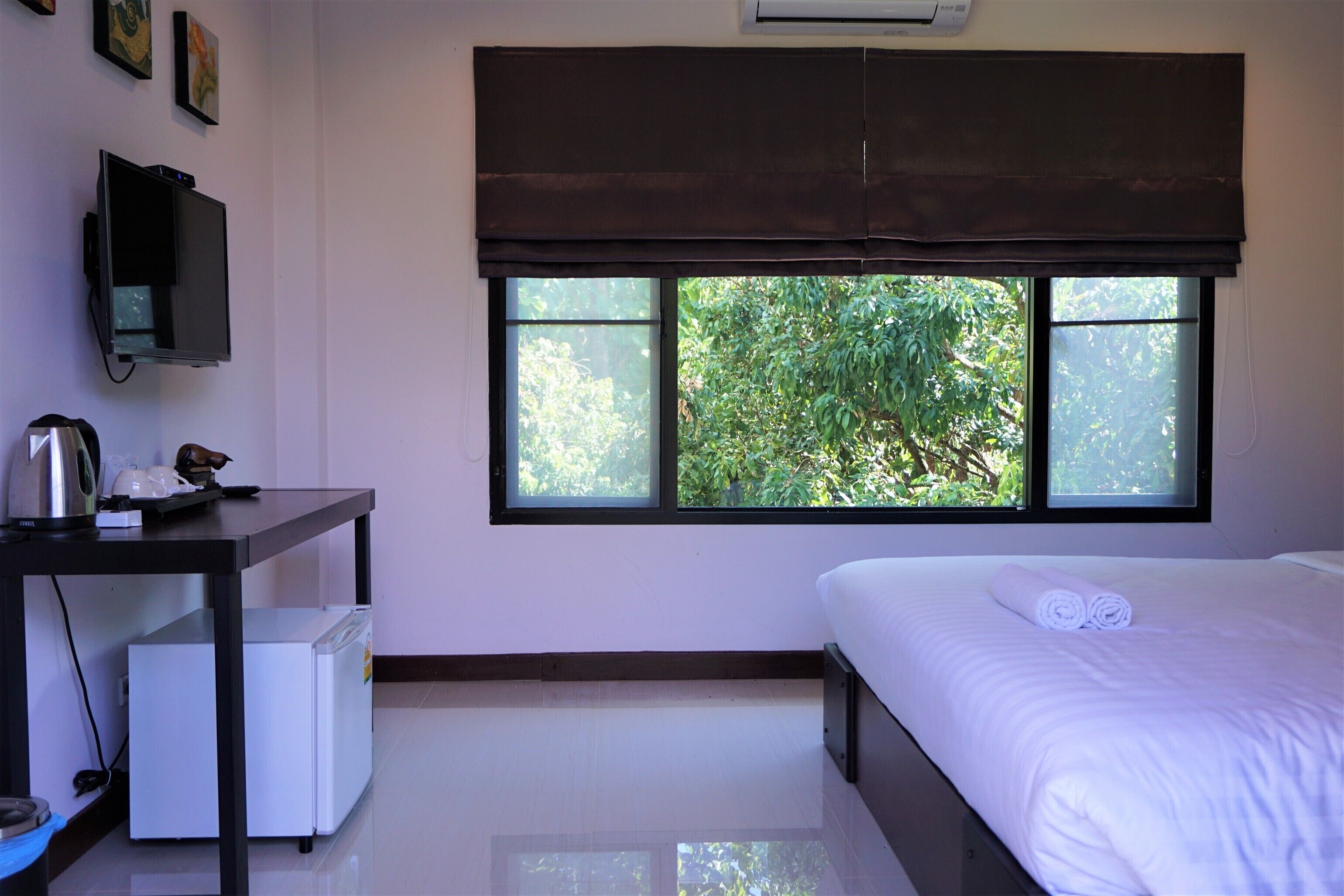 A hotel room with a bed, a desk with a computer, a small fridge, and a window with a view of greenery.
