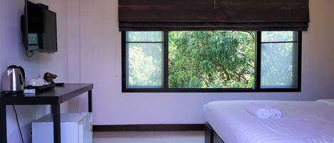 A hotel room with a bed, a desk with a computer, a small fridge, and a window with a view of greenery.