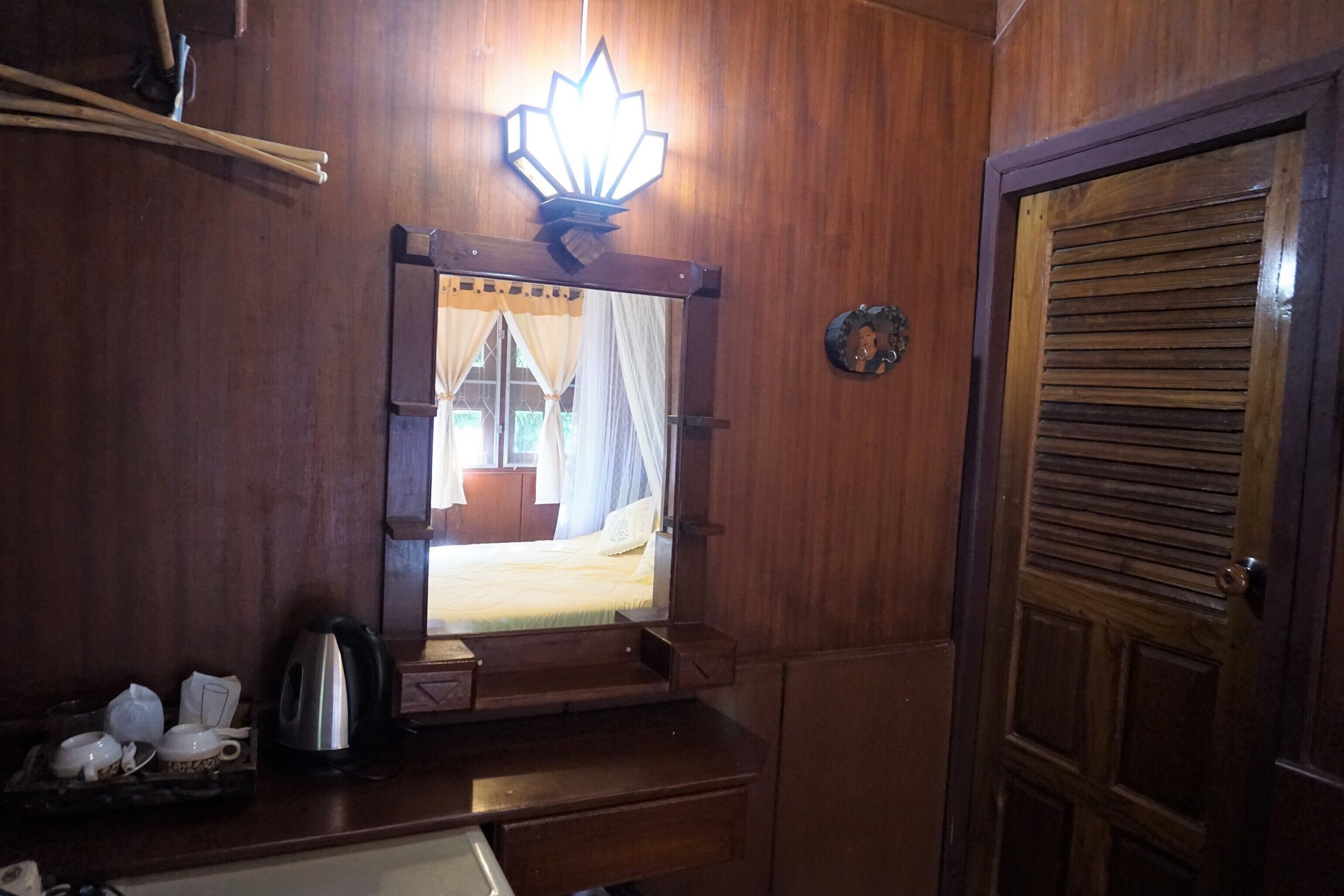 A wooden room with a bed, a nightstand, a kettle, and a mirror reflecting a window with curtains.