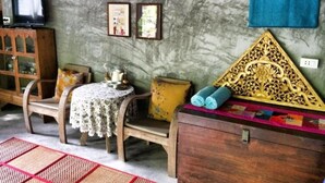 Une pièce comprenant un meuble en bois, une petite table recouverte d’une nappe en dentelle, une chaise, un coussin décoratif, un téléviseur fixé au mur et des tableaux encadrés.