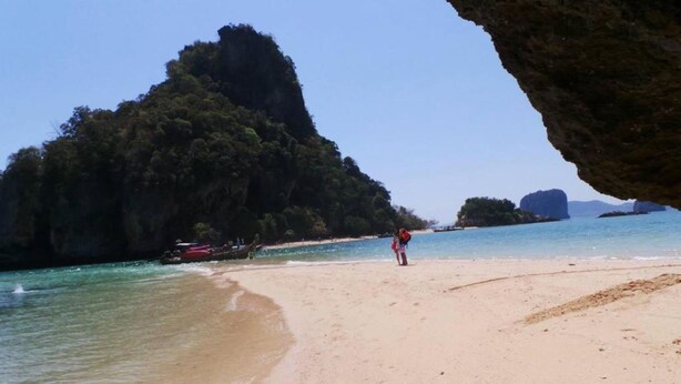 Une plage avec une eau turquoise et limpide, une grande formation rocheuse et une personne debout sur le sable.