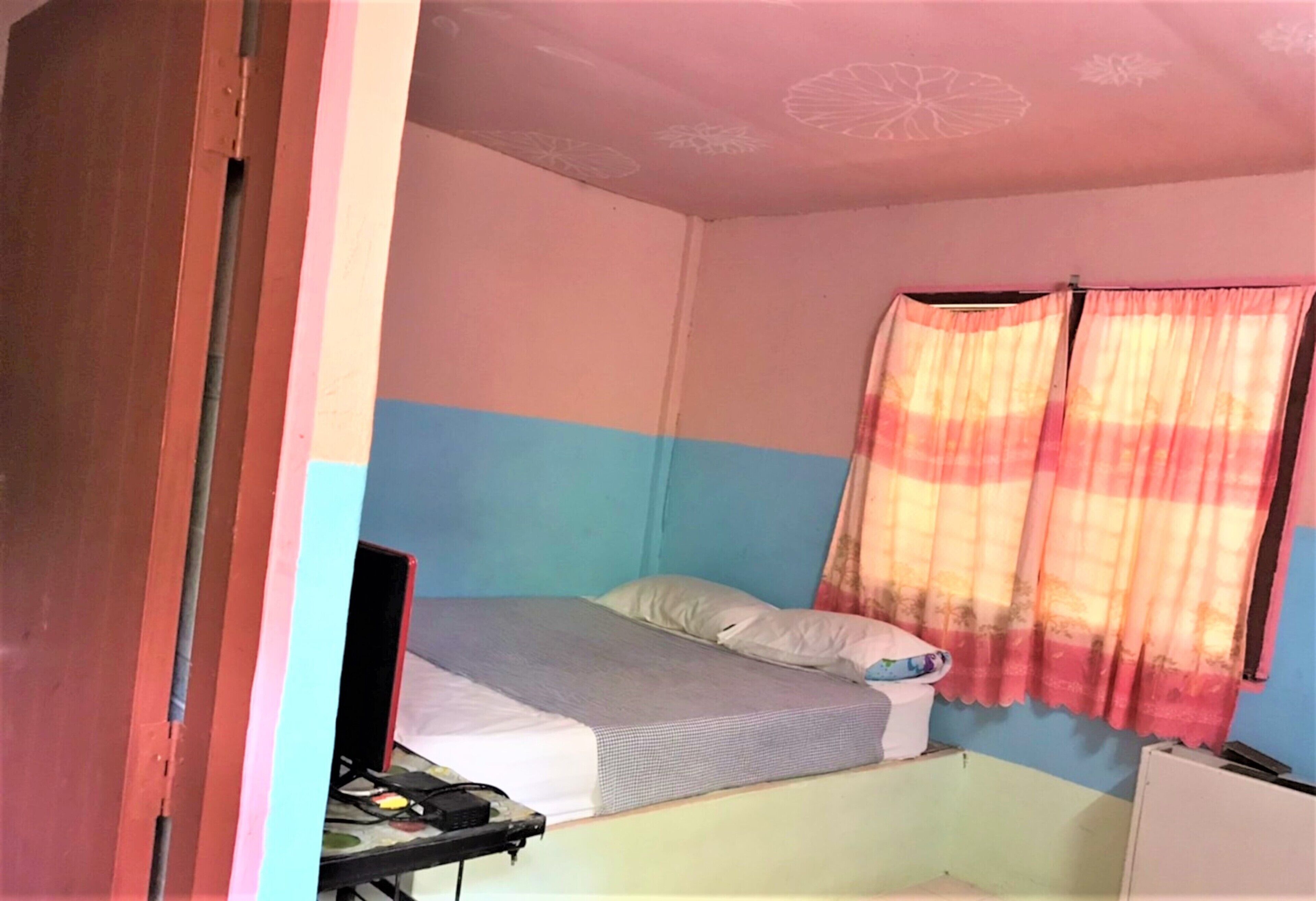 Una habitación pequeña con una cama individual, un escritorio y una silla, una ventana con cortinas, y una pared con estampado floral.