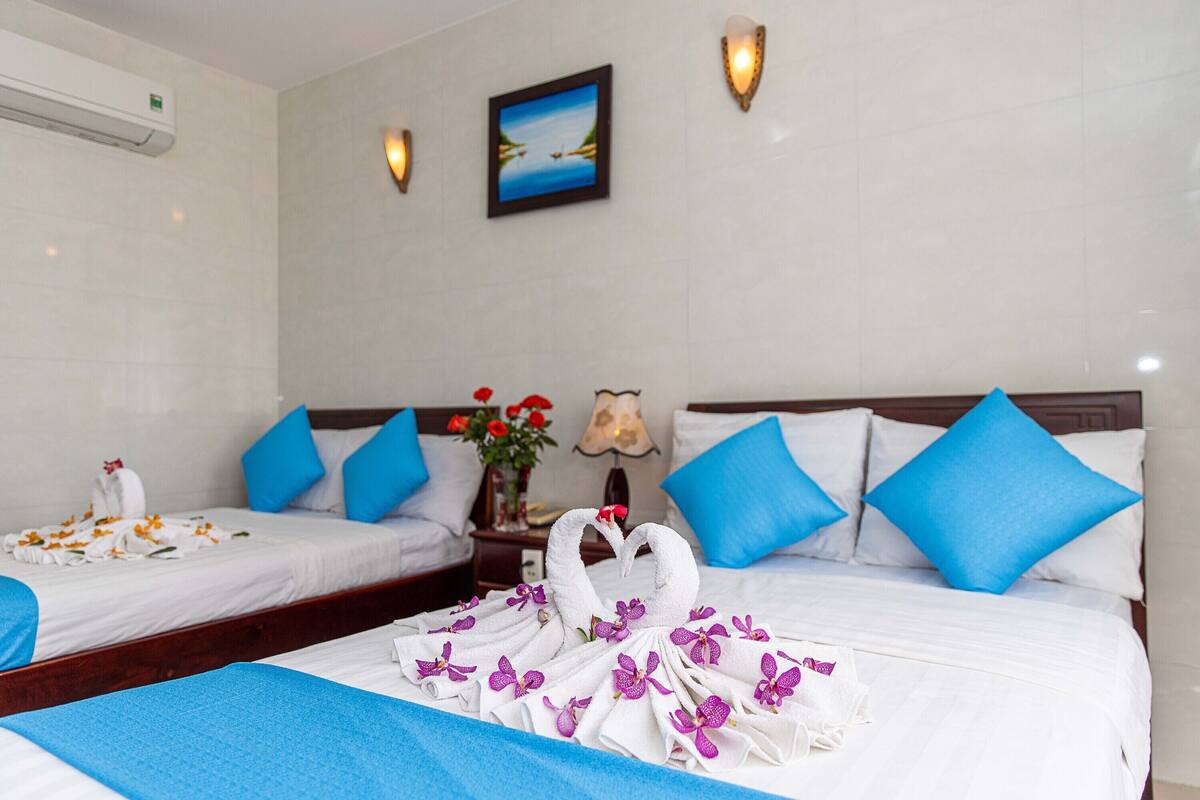 A hotel room with two beds, each with blue and white bedding, a wall-mounted air conditioner, a framed picture, and a nightstand with a lamp and flowers.