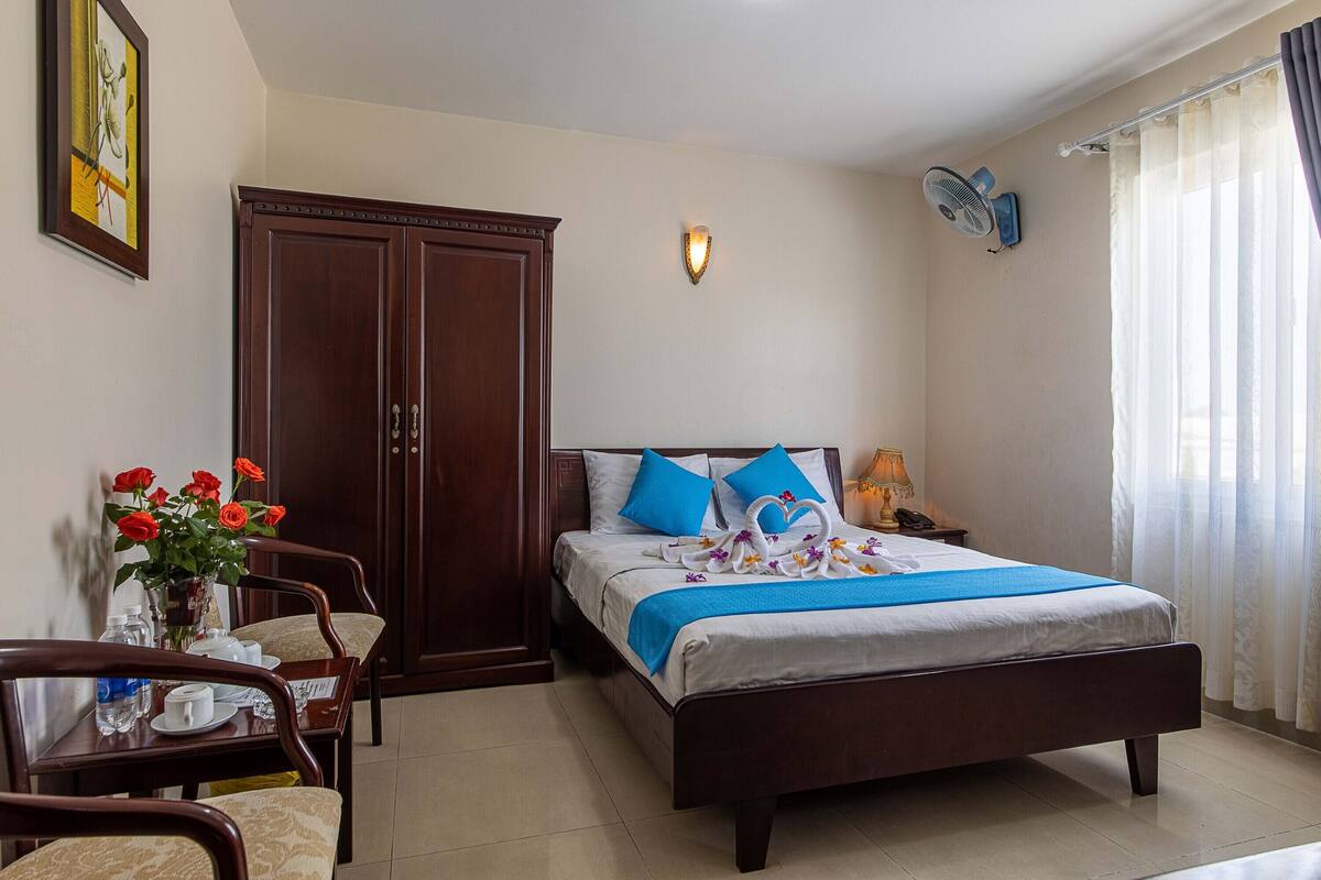 A neatly arranged hotel room with a bed, a wardrobe, a dining table with chairs, and a vase of flowers.