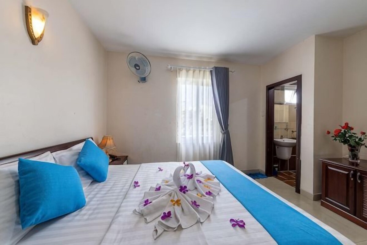 Habitación de hotel con una cama, una mesita de noche, un ventilador, una ventana con cortinas y un baño visible a través de una puerta abierta.