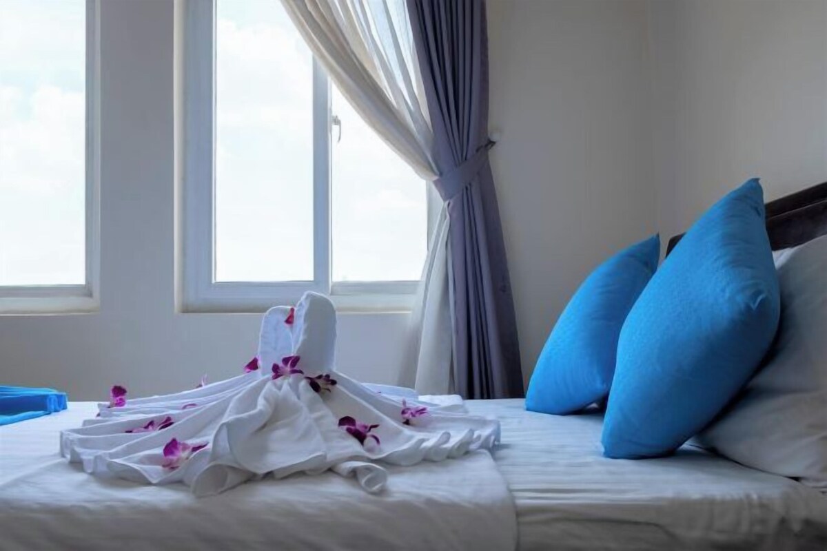 A hotel room with a bed, blue pillows, and a white towel with purple flowers.