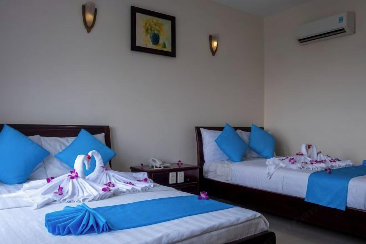 Habitación de hotel con dos camas, cada una con almohadas azules y una colcha blanca. Se observan cisnes decorativos hechos con toallas sobre las camas.