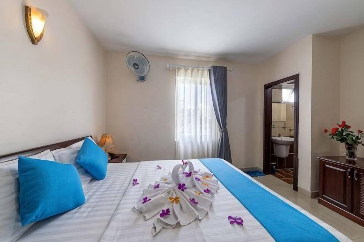 A hotel room with a bed, a bedside table, a fan, a window with curtains, and a bathroom visible through an open door.