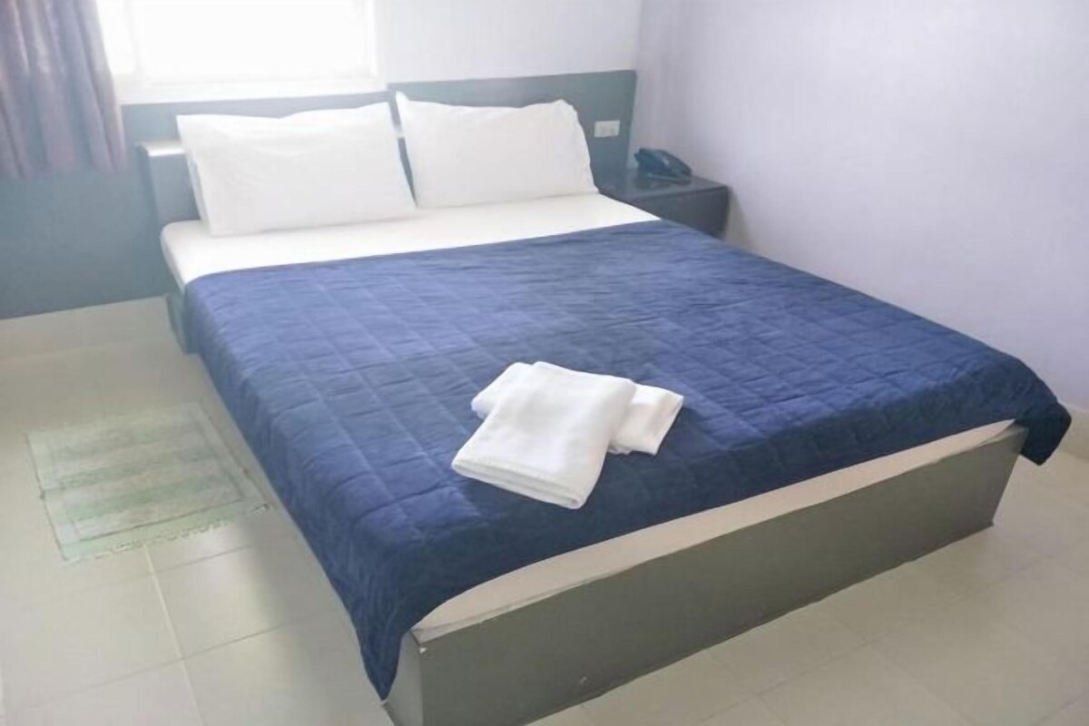 A neatly made bed with a blue bedspread and white pillows, a bedside table with a phone, and a window with curtains.