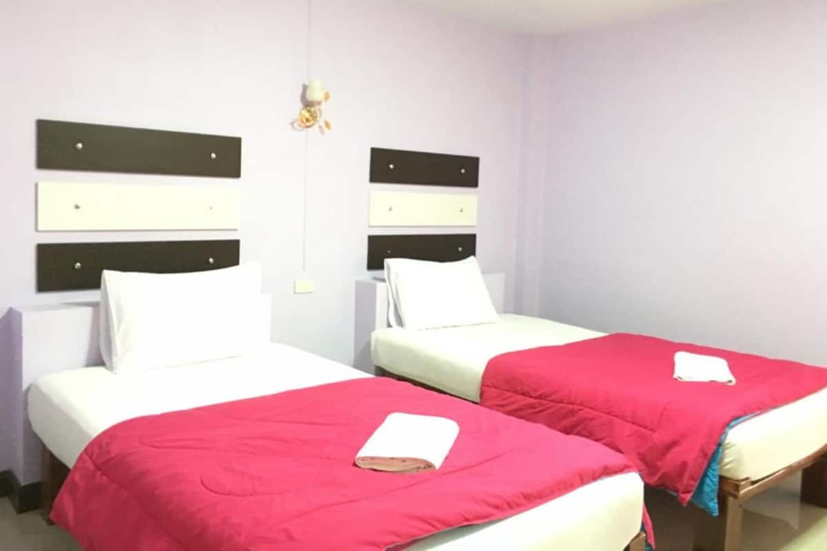 Une chambre d’hôtel avec deux lits, chacun doté d’un matelas blanc et d’une couverture rouge. Des tables de chevet sont fixées au mur, et elles sont surmontées de lampes blanches.