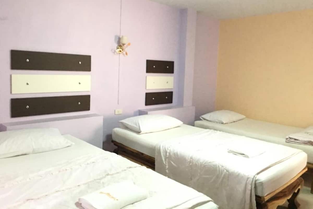 Une chambre d’hôtel avec trois lits simples, chacun recouvert d’une courtepointe et doté d’un oreiller blancs. Les murs sont peints dans des tons de violet pâle et de beige, et un petit objet décoratif est accroché au mur entre les lits.